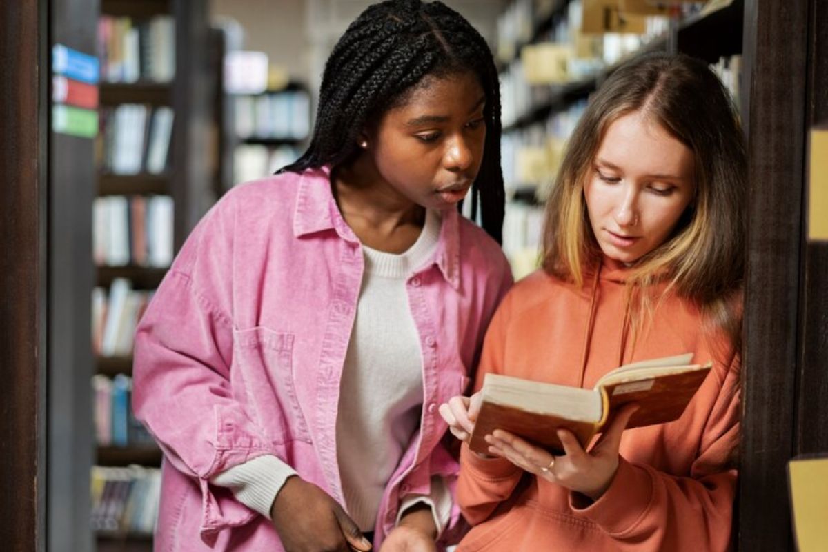 Duas alunas na biblioteca, uma está lendo o livro e outra olha por cima dos ombros