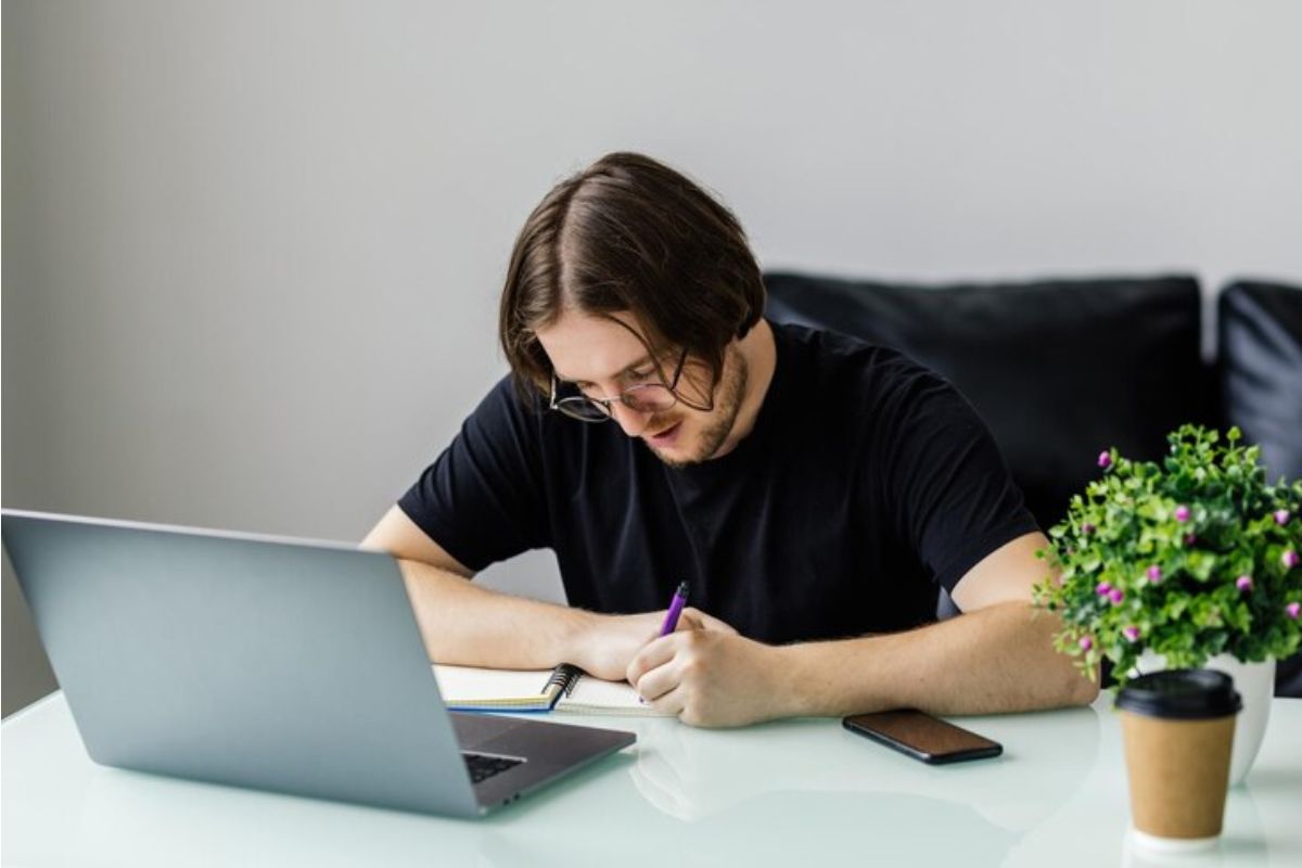 Um aluno faz anotações em seu caderno. Ele esta em casa, em frente ao seu notebook, onde pesquisa qual é o vestibular de medicina fácil de passar 2025