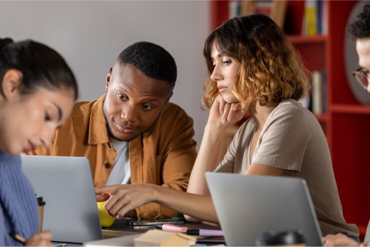 Três estudantes sentados em uma mesa. Uma colega ajuda o amigo a se inscrever no Vestibular UEMG 2025 apontado para seu notebook