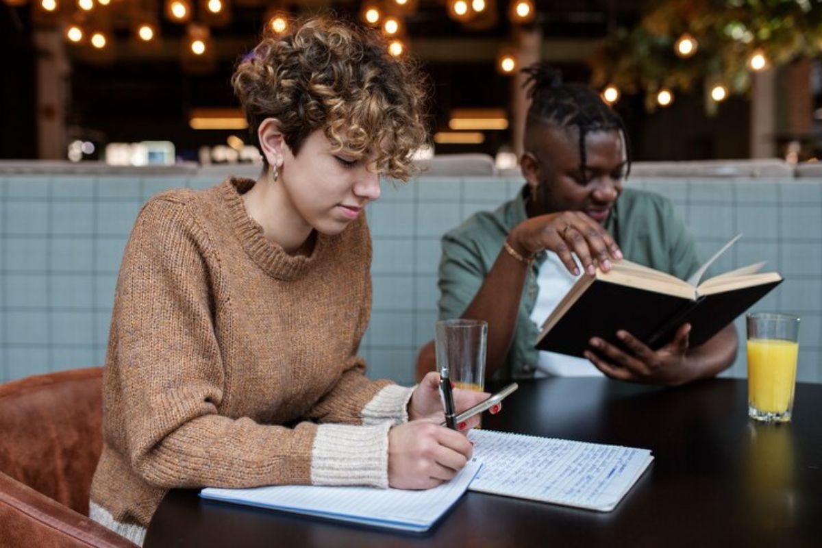 Dois estudantes estudam juntos em uma sala de aula. Um anota algo no caderno enquanto o outro lê o livro