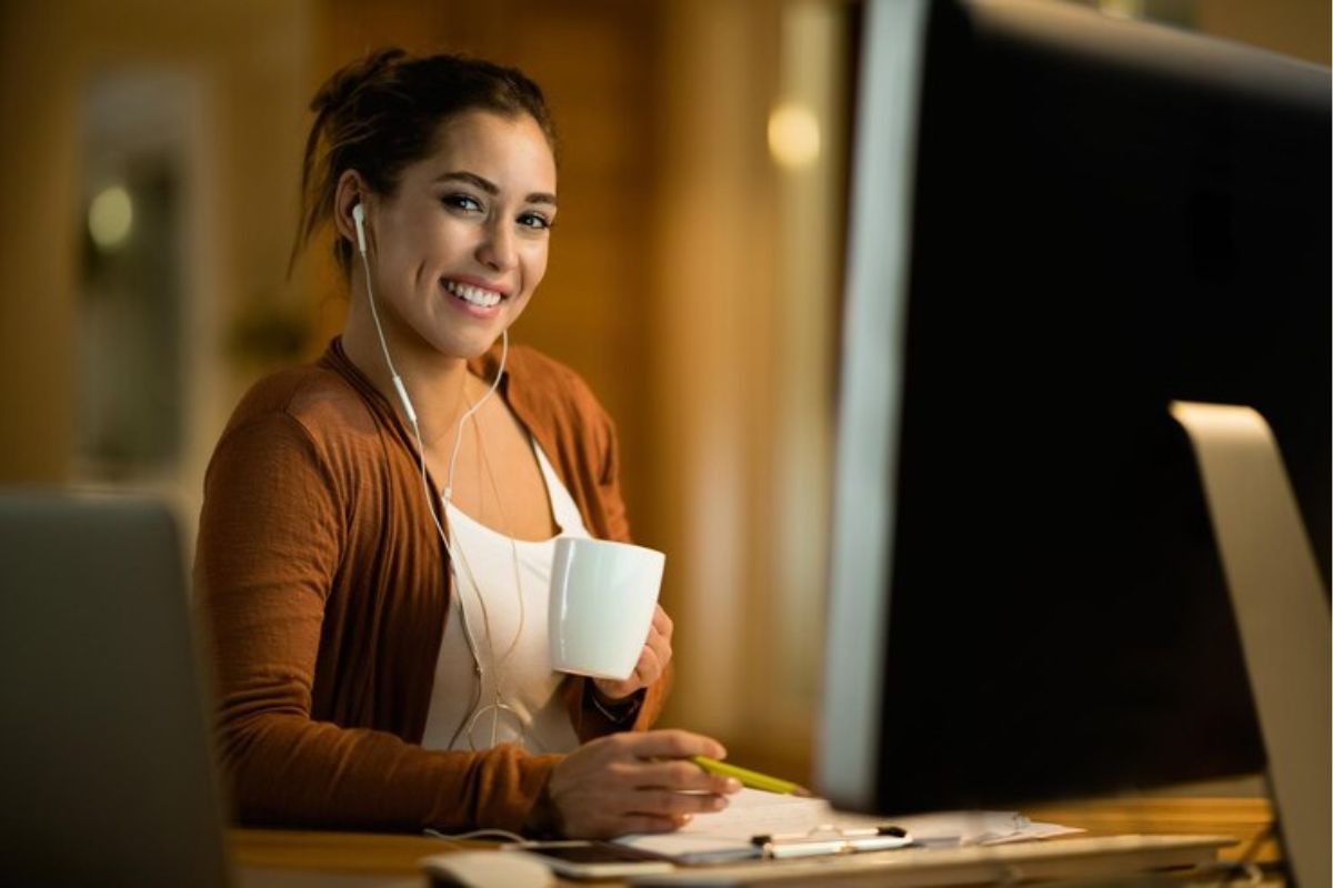 Uma estudante sorri para a foto. Ela está em seu quarto e estuda para o Vestibular UFMG 2025. Sentada em frente à mesa, onde está seu notebook, ela segura uma xícara de café