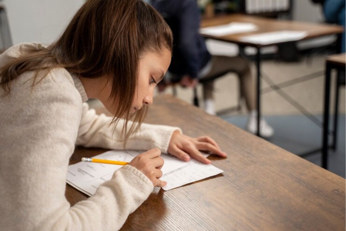 Uma menina na sala de aula. Apoiada na mesa, ela escreve em seu caderno