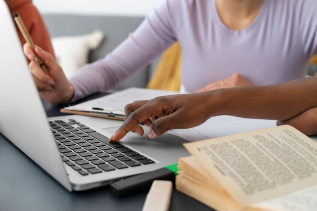 Uma estudante pesquisa em seu notebook, Na mesa ela também tem um livro aberto