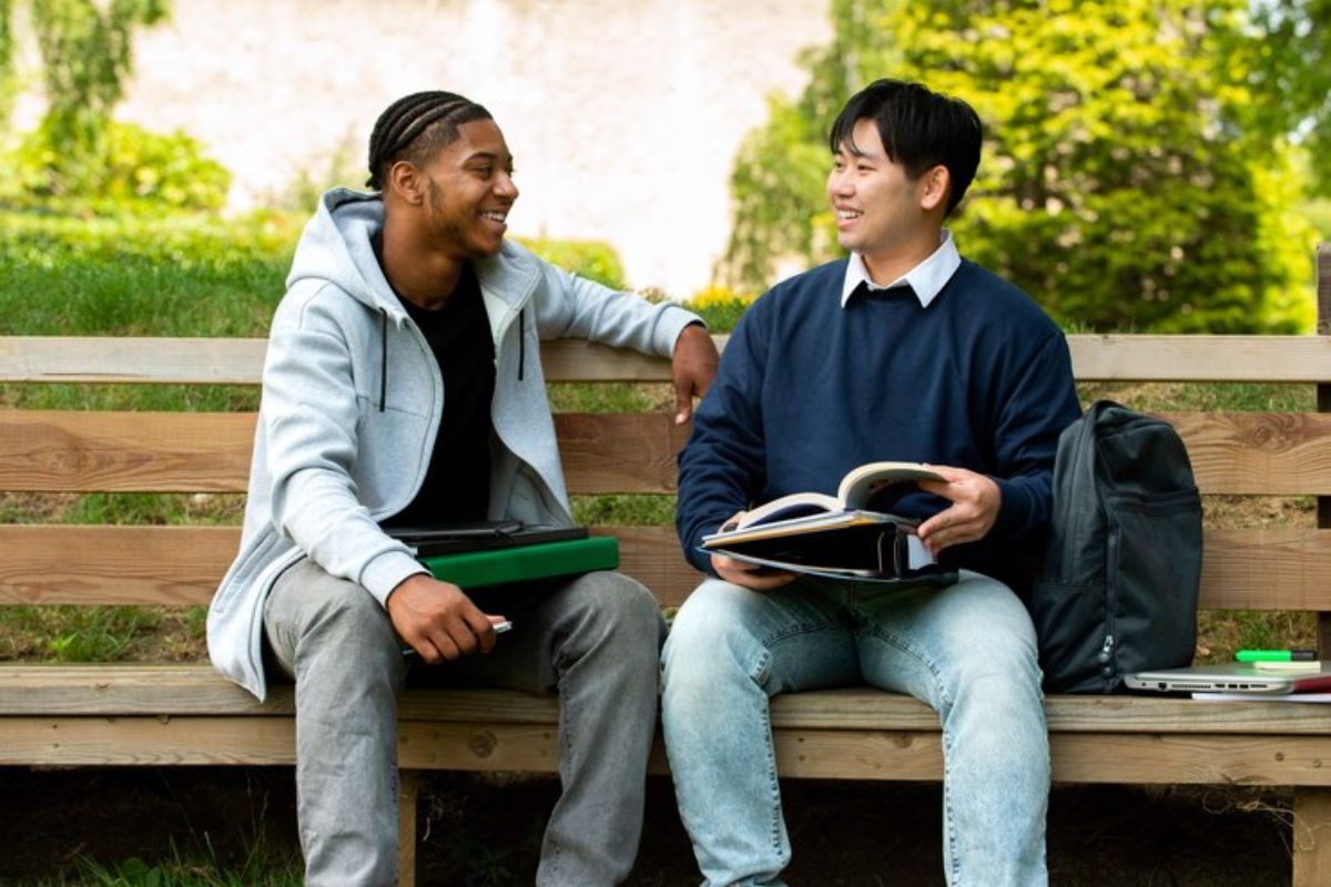 Dois estudantes estão sentados em um banco na praça do campus.