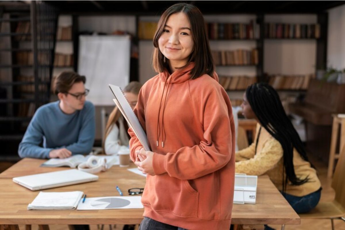 Uma estudante com seu material no colo olha para a foto. Ao fundo, há dois alunos sentados na mesa e conversam acerca do Vestibular UEG 2025