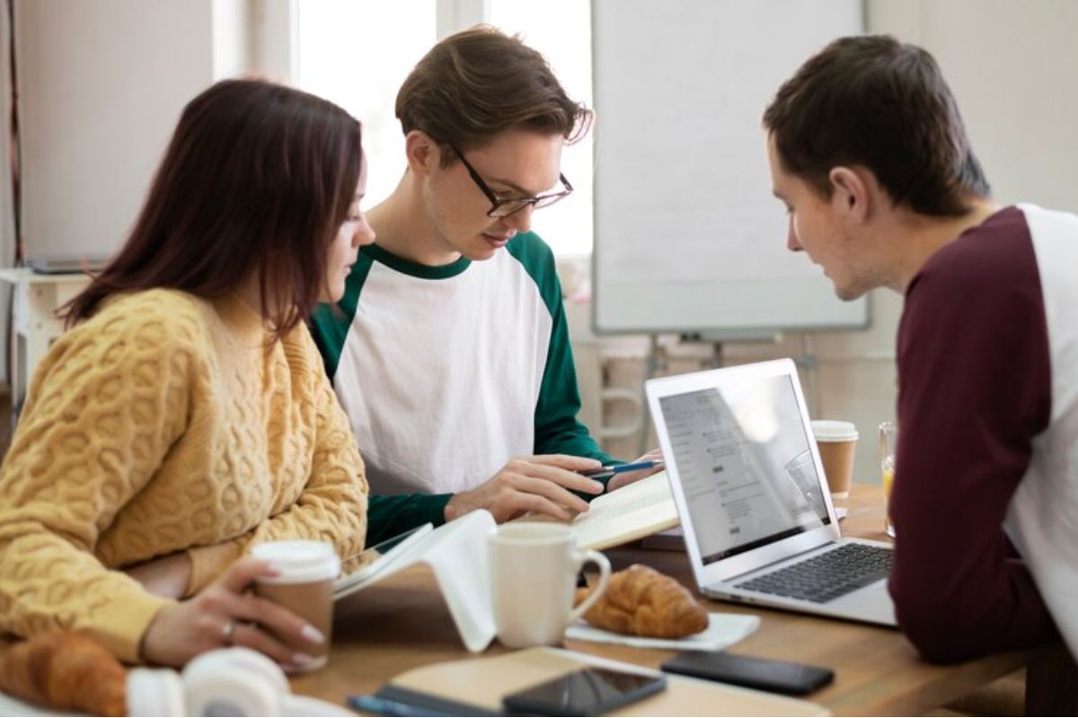 Três colegas conversam sentados em uma mesa com materiais escolares e um notebook ligado