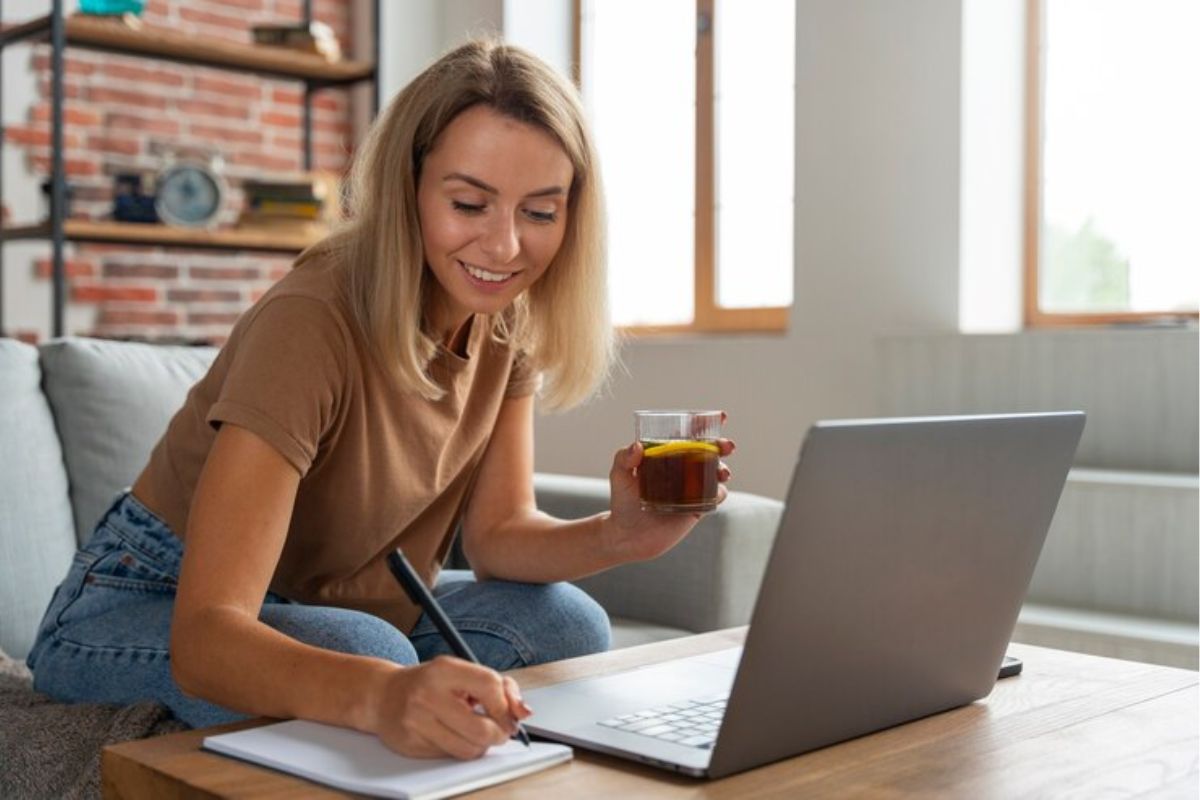 Uma estudante faz anotações em seu caderno sobre o Vestibular UFBA 2025 em frente ao seu notebook. Ela segura um copo de chá