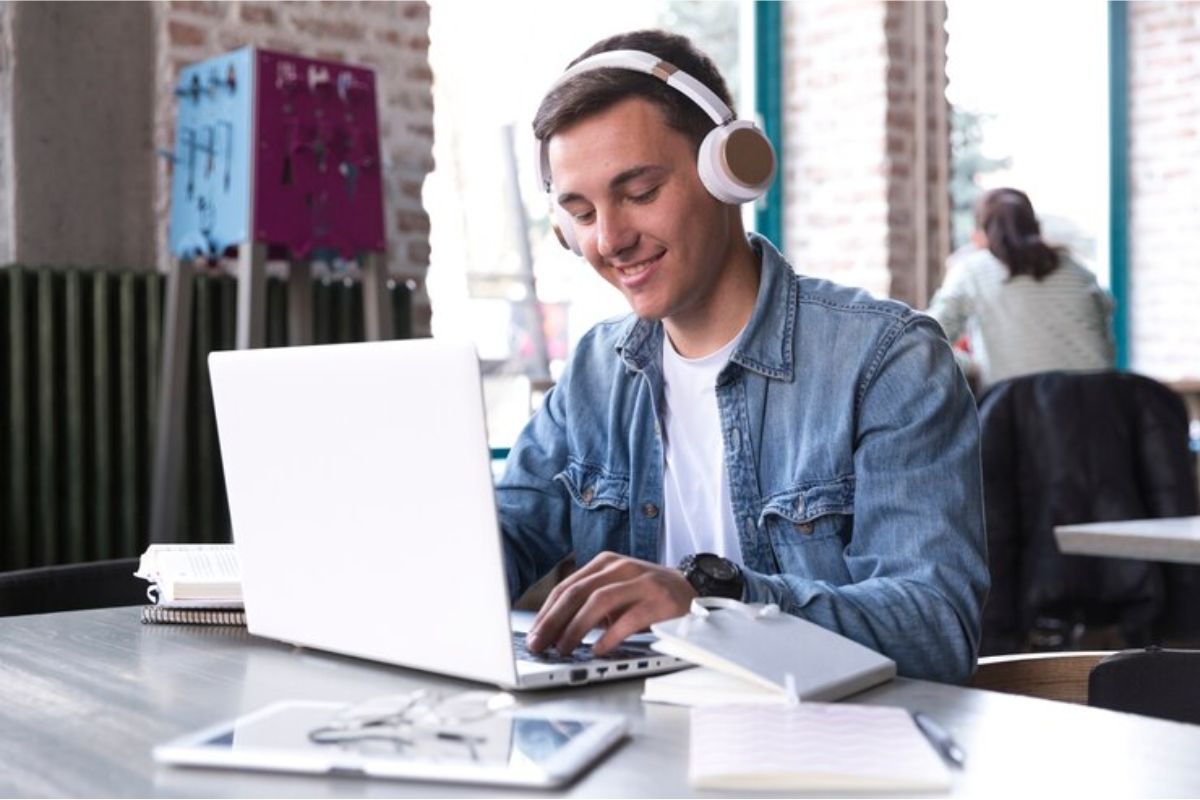 Um estudante com headphone mexe em seu notebook. Ele está em uma sala de aula da universidade 