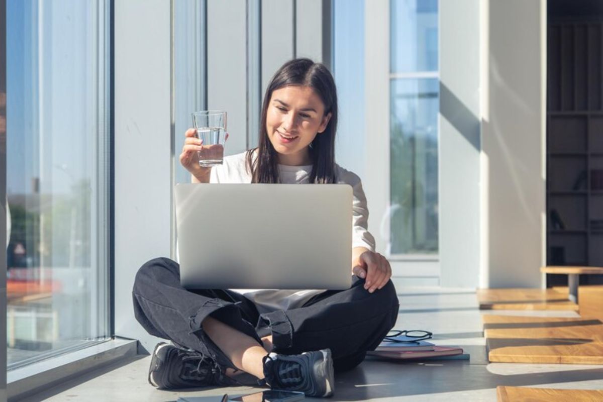 Uma menina sentada com o notebook no colo sentada na sala de casa ao lado da janela de vidro por onde entra o sol