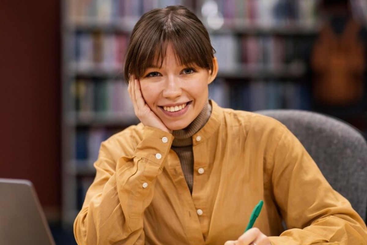 Uma estudante sorri para a foto. Ela apoia o cotovelo na mesa e sorri. Na sua frente está seu notebook e ela segura uma caneta