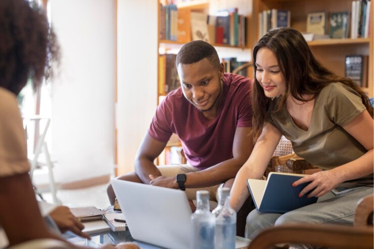 Três estudantes sentados em uma mesa. Uma colega ajuda o amigo a se inscrever no Vestibular UEMG 2025 apontado para seu notebook