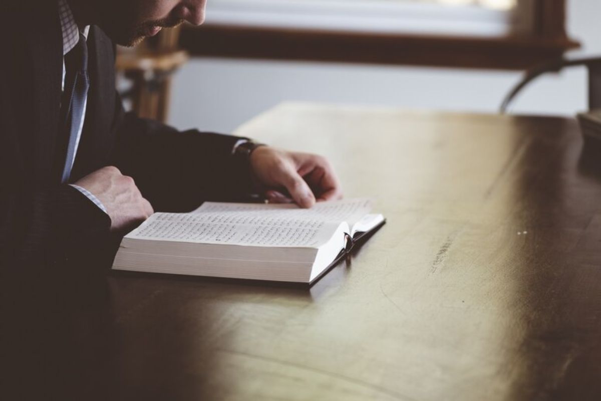 Uma pessoa com o livro aberto em cima da mesa, onde apoia os cotovelos enquanto lê