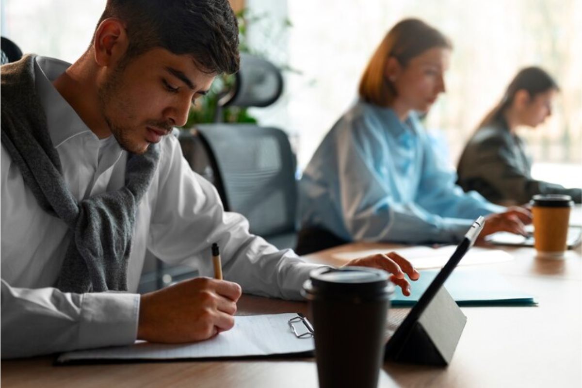 Um estudante na sala de informática usa o notebook. Ele faz anotações em seu caderno acerca do Vestibular Unifesp 2025