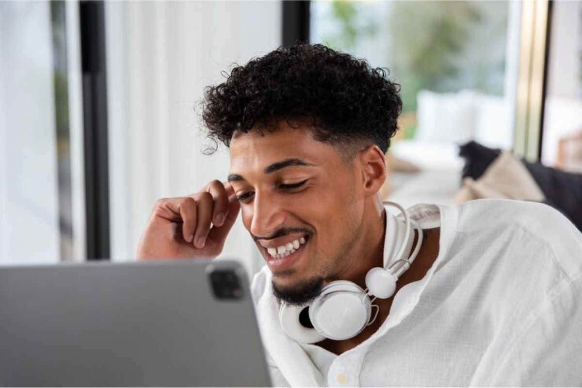 Um estudante sorri em frente ao seu notebook. Há um headphone que passa pelo seu pescoço