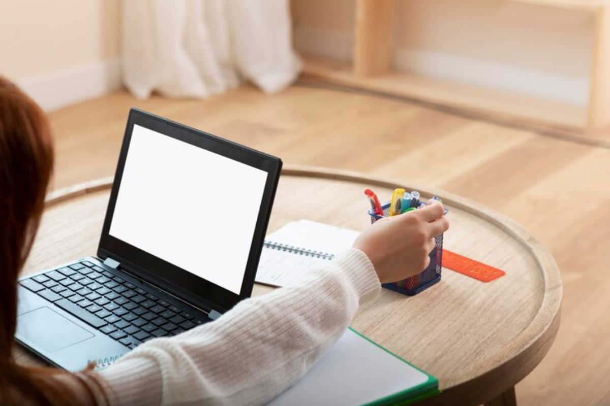 Uma estudante verifica o edital do Vestibular UEM 2025 em seu notebook que está em cima de uma mesa redonda. A estudante pega uma caneta colorida que está em uma cestinha na mesa 