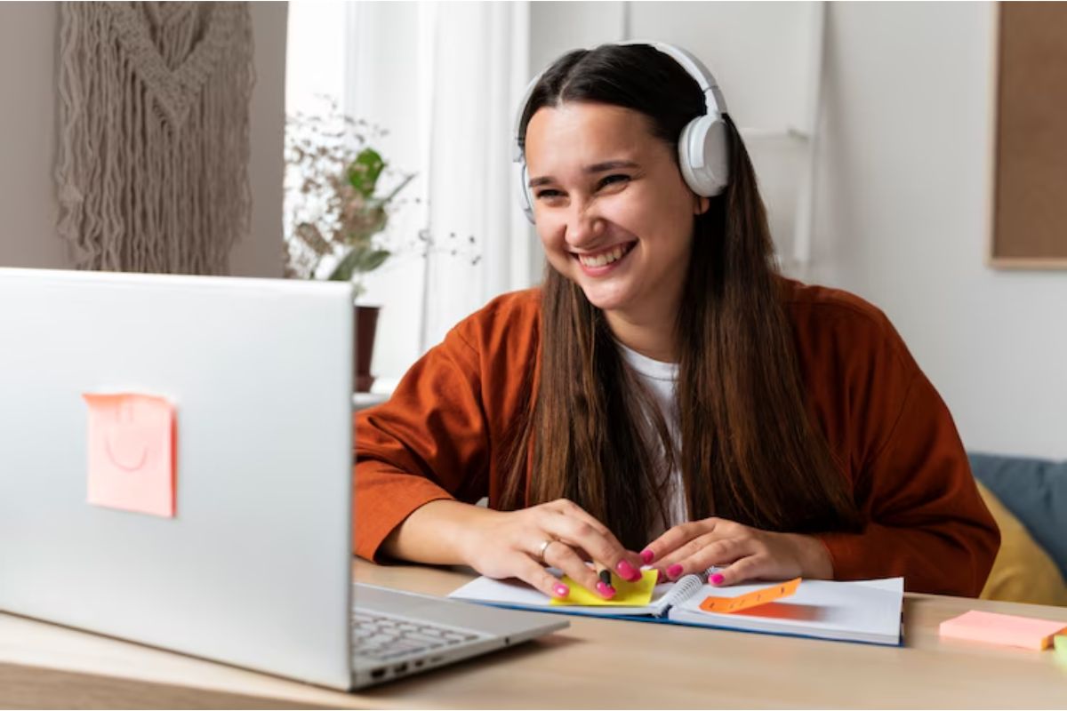 Uma mulher em frente ao seu notebook estudando. Ela sorri com um headphone na cabeça