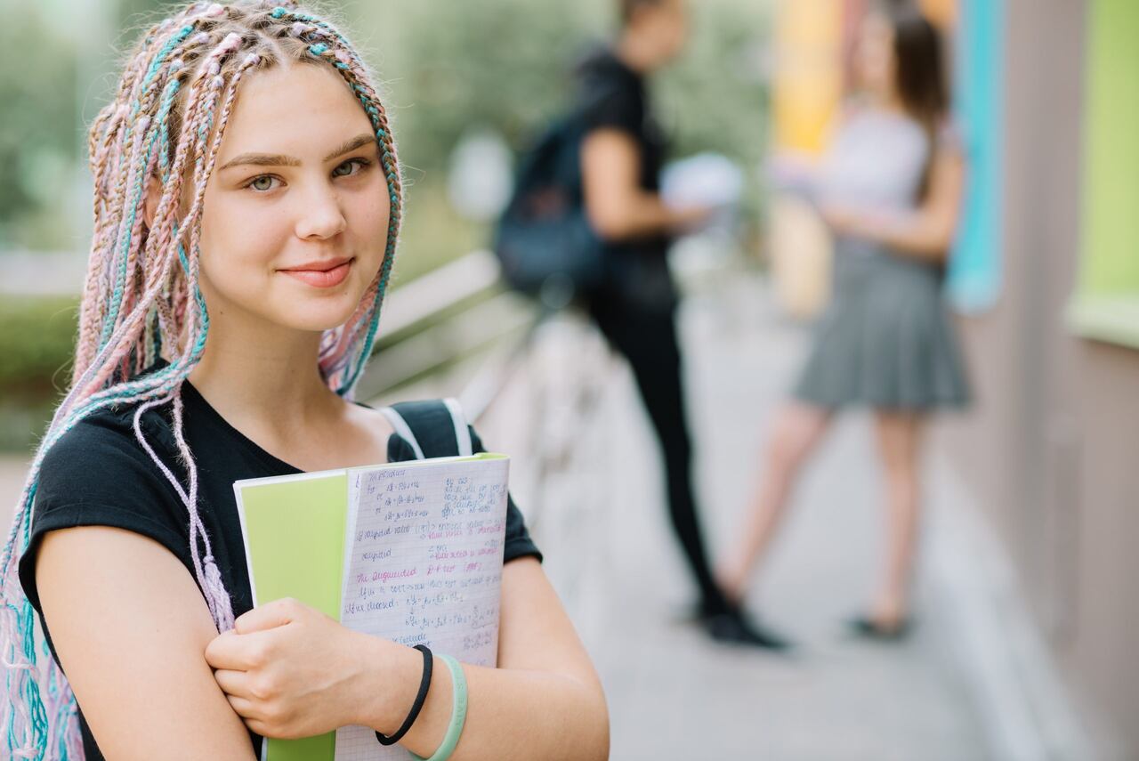 Jovem segurando livro no campus da universidade com colegas ao fundo 