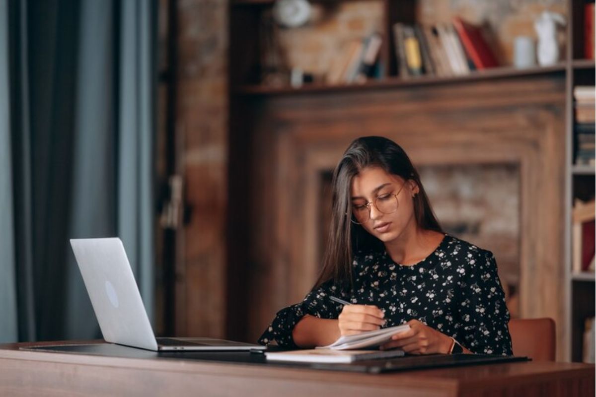 Uma menina em frente ao notebook, sentada em sua sala, faz anotações em seu caderno sobre o Vestibular UEMA 2025