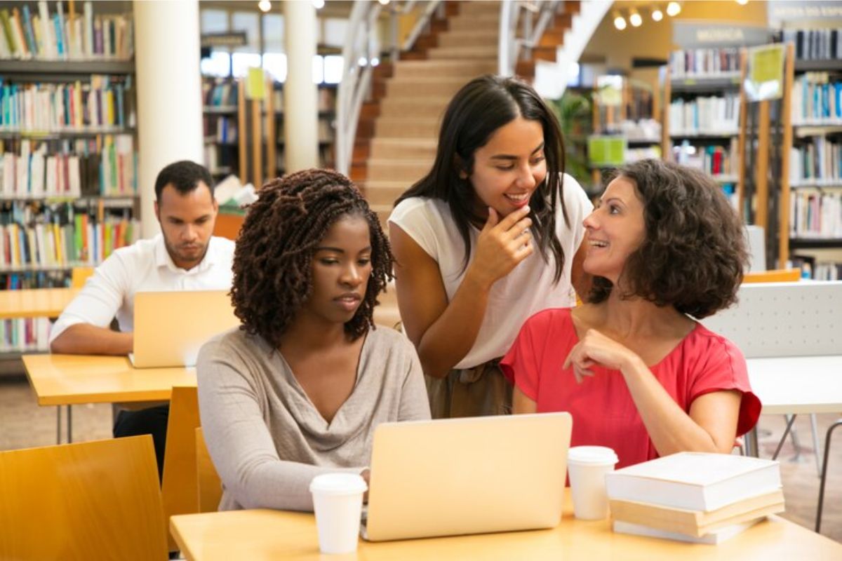 Um trio de amigas na biblioteca. Uma aluna usa o notebook para acessar informações sobre o Caixa FIES 2025. Ao fundo outro aluno está sentado e usa seu notebook sozinho na mesa