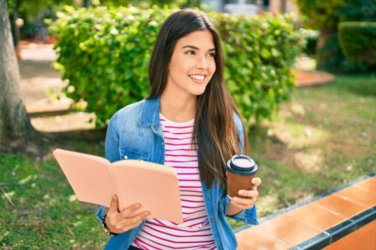 Uma estudante com um caderno aberto e um copo de café na mão, ela sorri ao ar livre