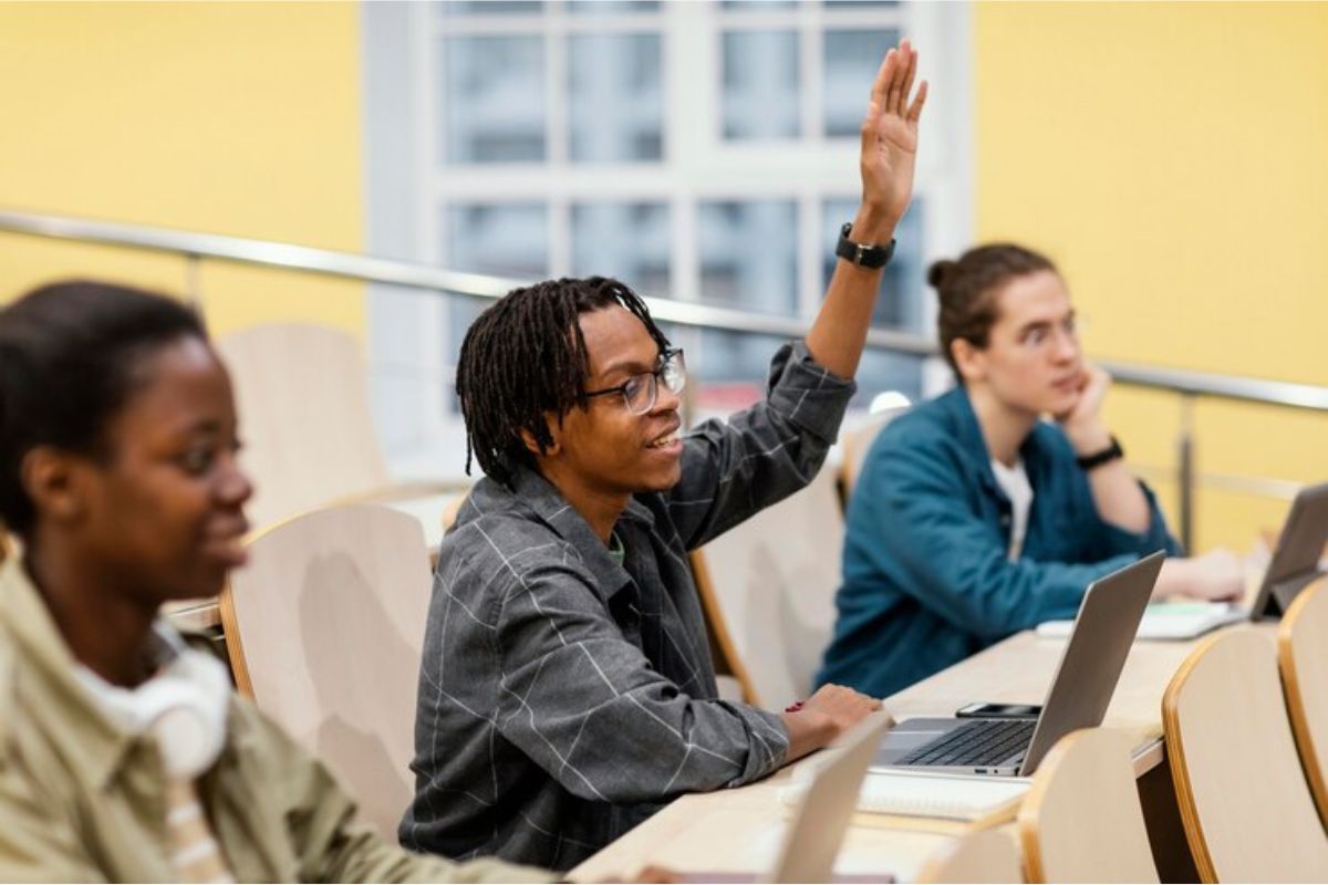 Um estudante na sala de aula levantando a mão. Ela está entre dois colegas 