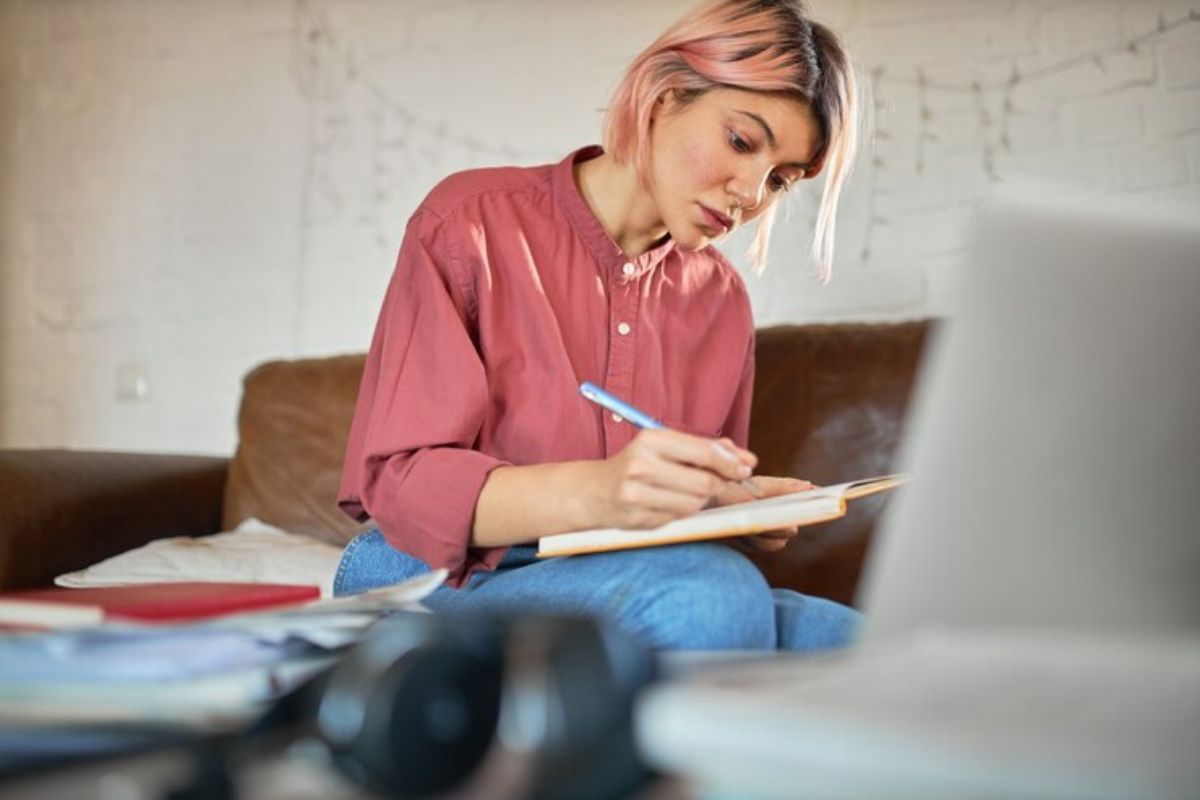 Uma aluna estudando para a Prova da USP 2025. Ela está sentada escrevendo em seu caderno vendo o vídeo que passa no notebook