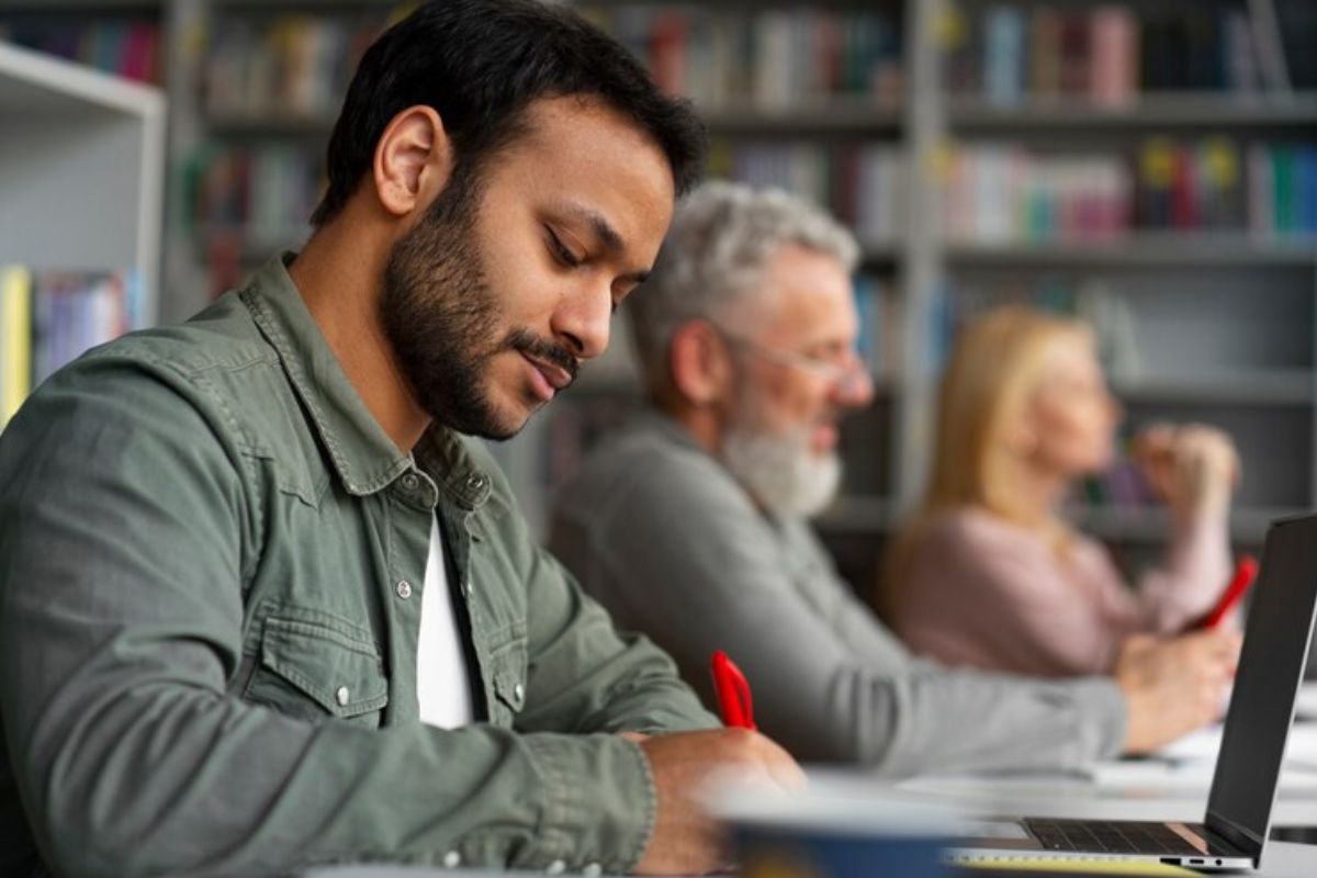 Três alunos na aula (mais velhos). Eles fazem anotações no caderno. Um está com o notebook em cima da mesa 