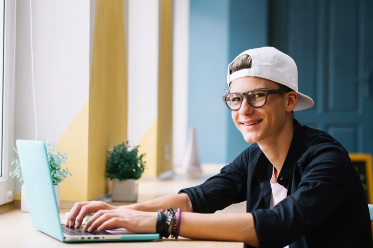 Um aluno de boné branco olha para a foto enquanto digita em seu notebook