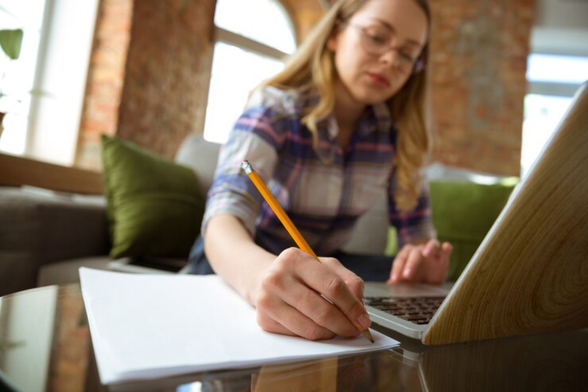 Uma aluna estudando para a Prova da USP 2025. Ela está sentada escrevendo em seu caderno vendo o vídeo que passa no notebook