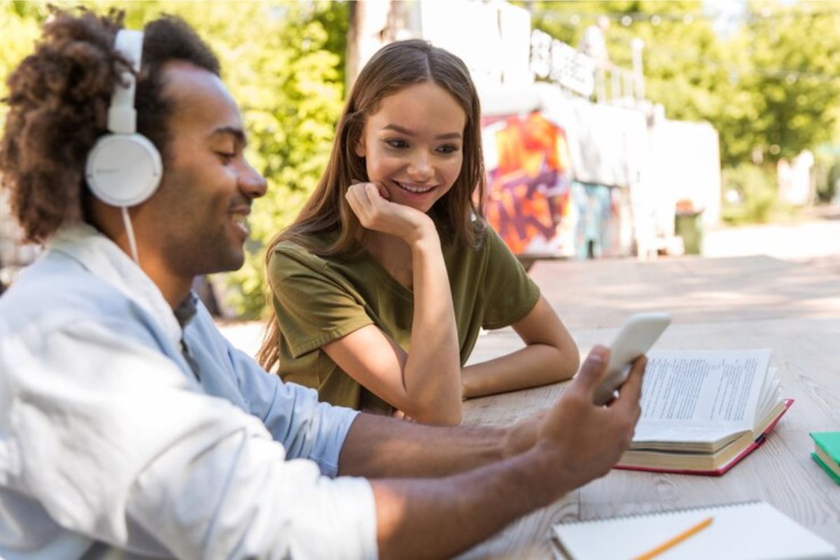 Dois estudantes ao ar livre na faculdade, conversam após o Vestibular UERJ 2025. O amigo mostra algo no celular para a colega que olha entusiasmada 