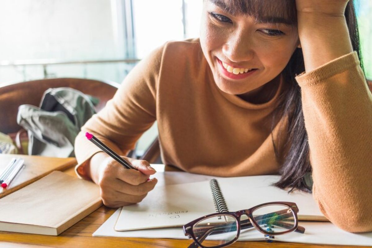 Uma estudante escrevendo em seu caderno. Ela estuda para o Vestibular Unimontes 2025. Na mesa é possível ver um óculos além dos seus materiais 