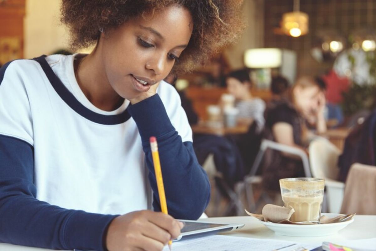 Uma estudante escrevendo em seu caderno com um lápis. Ela está sentada na sala de aula