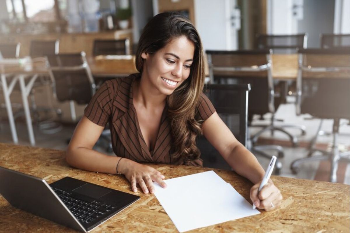 Uma estudante escrevendo e uma folha. Ela estuda para o Vestibular Unimontes 2025. Na mesa está o seu notebook