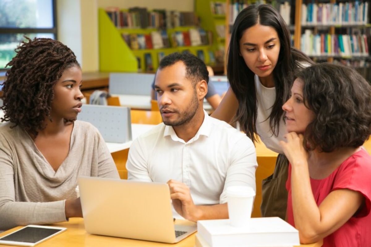 Um grupo de quatro alunos na biblioteca. Eles se reúnem em frente a um notebook, onde conferem o FIES Mestrado e Doutorado 2025