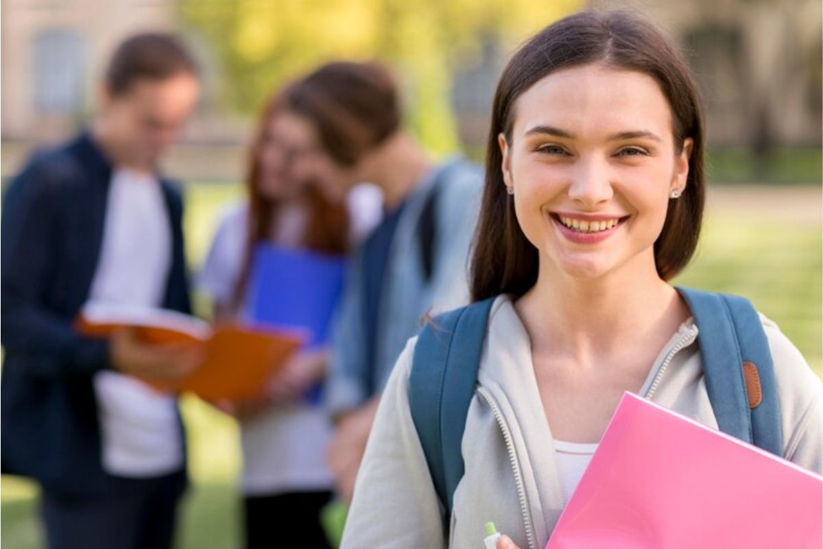 uma estudante segura um caderno rosa e sorri para a foto. Ao fundo há dois colegas conversando sobre o FIES UniFG 2025