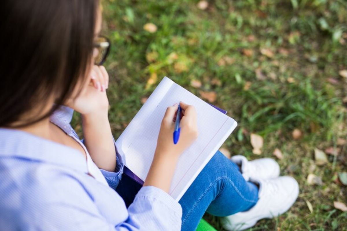 Uma menina faz anotações no caderno. Ela está sentada no gramado da escola