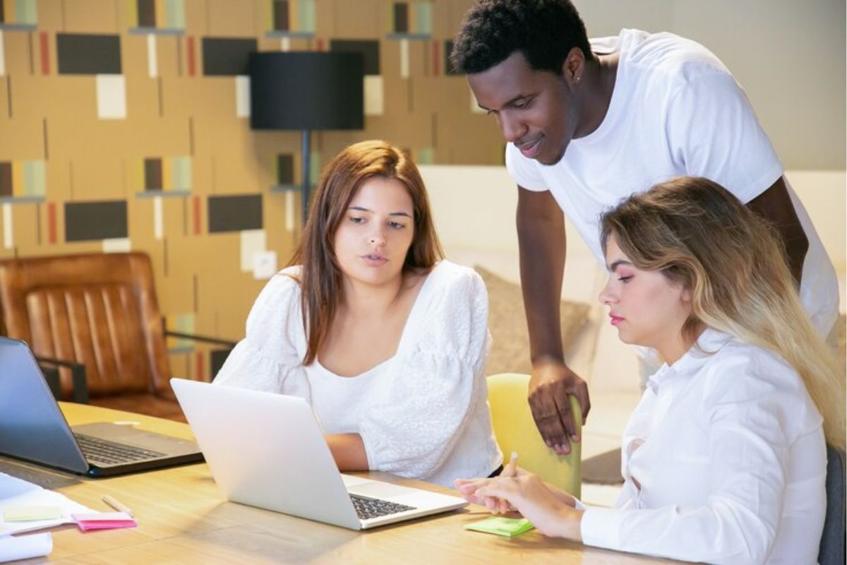 Três estudantes na biblioteca. Uma colega em frente ao notebook mostra o site do SisFies 2025