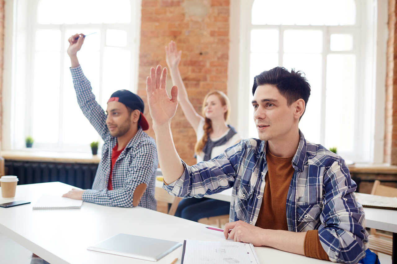 Universitários em sala de aula levantam a mão para responder questão do professor 