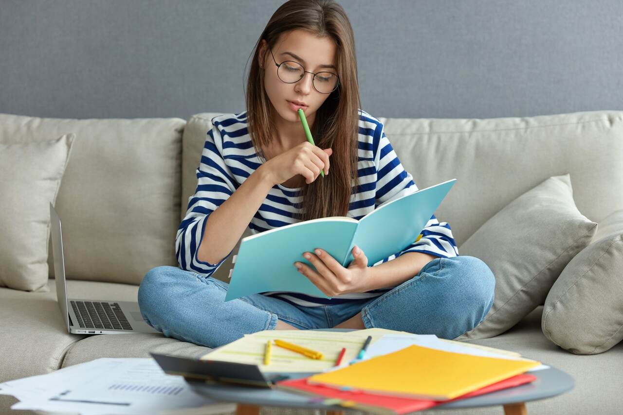 Adolescente estuda concentrada conteúdo de livro em sua mão sentada no sofá diante livros e cadernos 